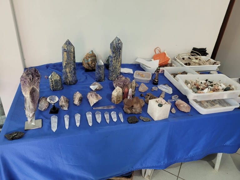 Uma mesa coberta com um tecido azul exibe diversos cristais e pedras, incluindo cristais de quartzo, amostras de outras rochas e utensílios associados à mineração.