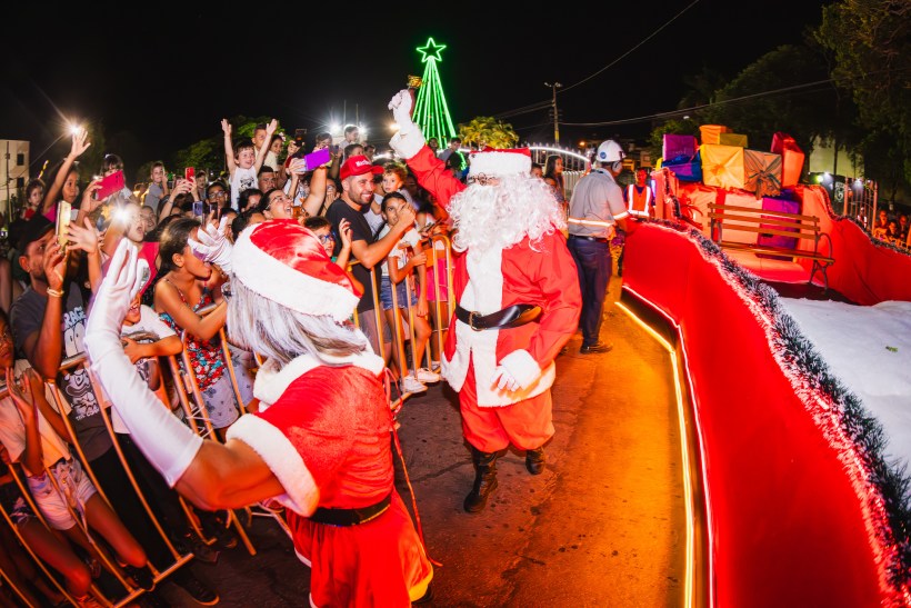 Celebração natalina com Papai Noel e uma multidão animada em um desfile festivo, com decoração iluminada e árvore de Natal ao fundo.