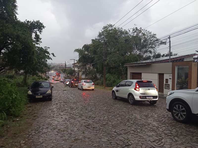 Rua em Divinópolis com carros estacionados e tráfego em dia chuvoso, evidenciando condições meteorológicas adversas.