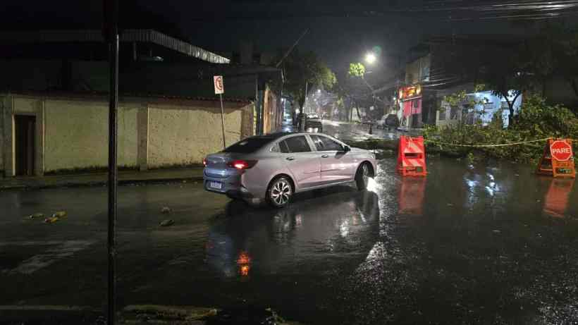 Um carro prata em uma rua alagada à noite, com galhos de árvores caídos e sinalização de interdição visível ao fundo.