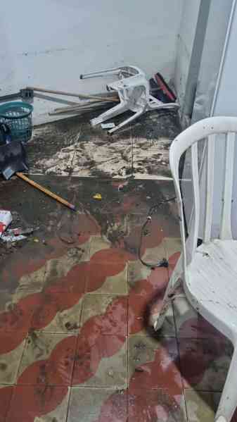 Interior de um espaço com piso sujo e entulho, incluindo uma cadeira branca de plástico e ferramentas espalhadas, indicando danos causados por alagamento.