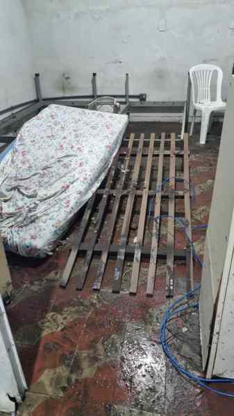 Interior de uma residência com piso molhado e madeiramento danificado, mostrando um colchão e um estrado de cama, além de uma cadeira plástica.
