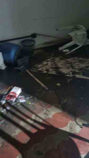 Interior de uma casa mostrando danos causados por alagamento, com móveis deslocados e sujeira no chão.