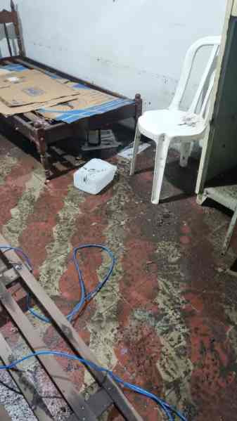 Interior de um quarto com piso molhado e objetos desorganizados, incluindo uma cadeira branca e uma cama com papelão em cima.