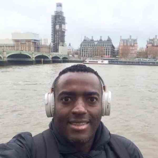 Um homem sorridente usando fones de ouvido tira uma selfie à beira do rio, com o Parlamento britânico e o Big Ben ao fundo, parcialmente coberto por andaimes.