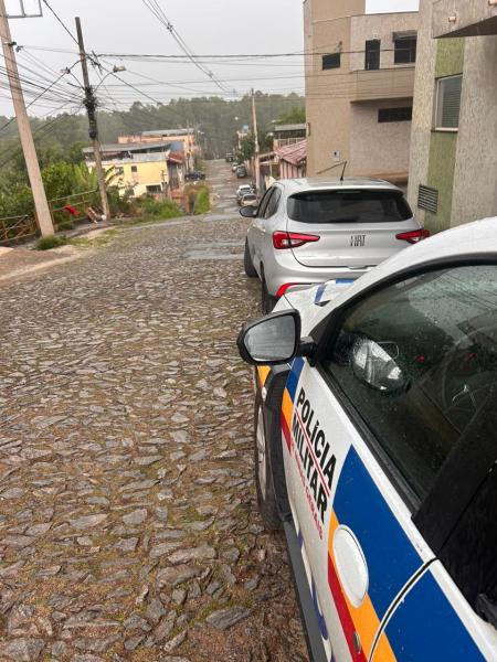 Imagem de uma rua de paralelepípedo em uma área urbana, com um carro da Polícia Militar em primeiro plano e outros veículos estacionados ao fundo, em um dia chuvoso.