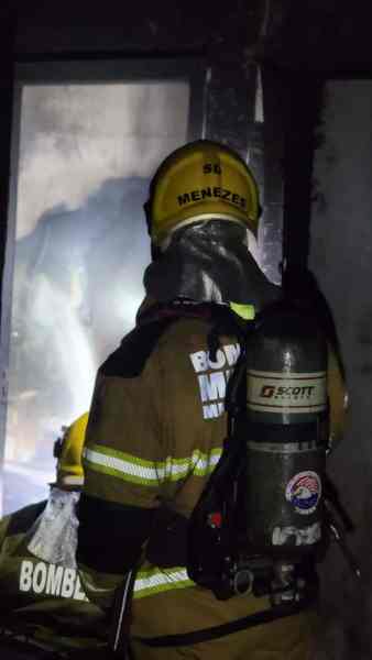 Bombeiros em ação dentro de uma residência em chamas, usando equipamento de proteção e respiradores, com fumaça visível ao fundo.