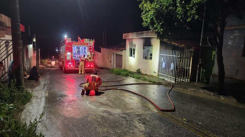 Veículo de bombeiros estacionado em uma rua durante a noite, com mangueiras estendidas no chão e equipe trabalhando na próxima residência, que apresenta danos visíveis.