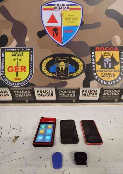 Equipamentos eletrônicos apreendidos pela polícia, incluindo dois celulares e um dispositivo codificador, em frente a um fundo com emblemas da Polícia Militar.
