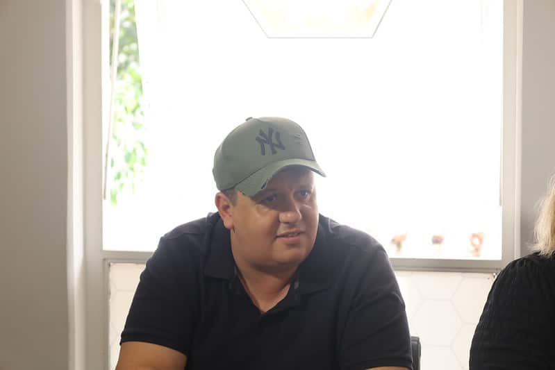 Homem com boné sentado em mesa durante reunião, com janelas ao fundo.