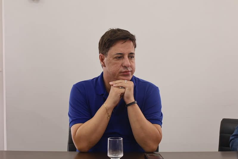 Homem com camisa azul, posicionado pensativo em uma mesa, com um copo de água à sua frente.