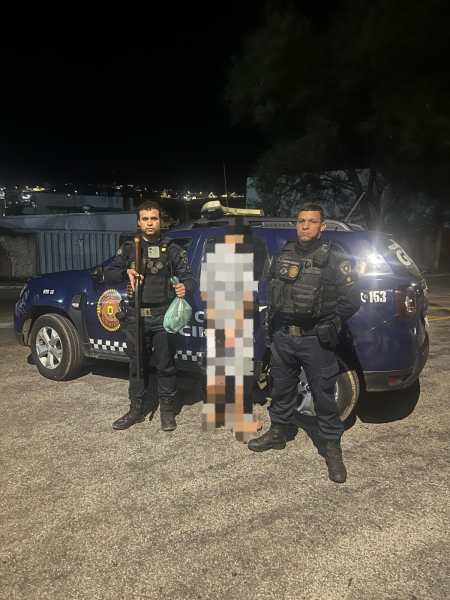 Dois agentes da Guarda Civil Municipal de Nova Serrana posam ao lado de um indivíduo suspeito detido, em frente a um veículo da corporação. Um dos agentes segura um bastão, enquanto o outro segura um invólucro. A cena ocorre à noite.