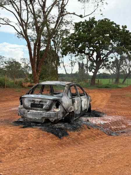 Veículo Ford Fiesta Sedan completamente queimado em estrada rural, com árvores ao fundo e solo de terra vermelha.