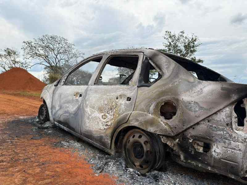 Veículo Ford Fiesta Sedan completamente queimado, com danos visíveis e rodas danificadas, localizado em uma estrada na zona rural.