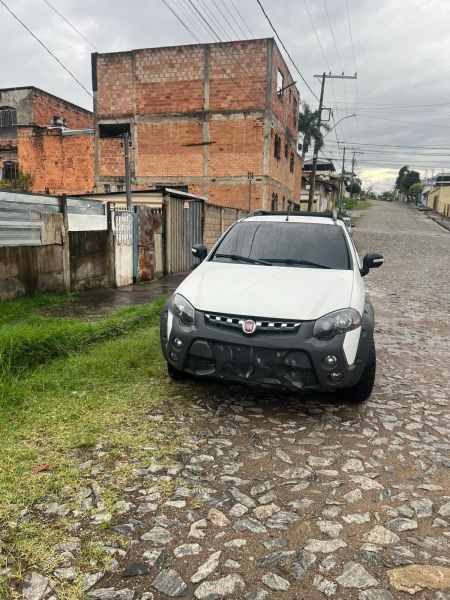 Um veículo Fiat Argo branco estacionado em uma rua de paralelepípedo, com edificações de tijolos ao fundo e céu nublado.