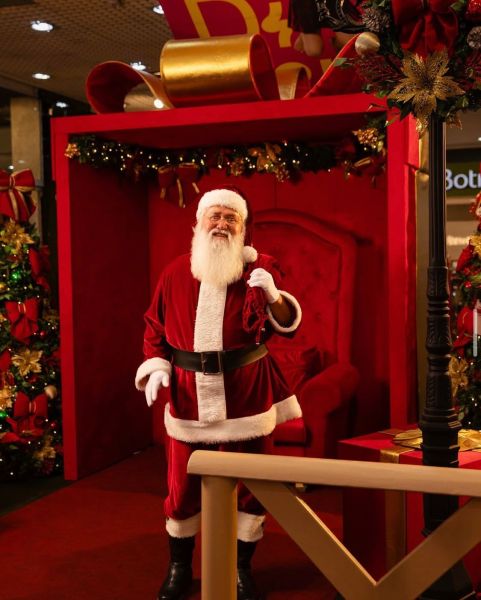 Um Papai Noel em um cenário festivo com decoração de Natal, posando em frente a uma cadeira vermelha decorada com laços e presentes.