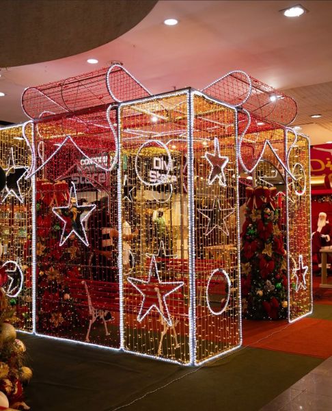 Decoração de Natal iluminada em um centro comercial, apresentando uma estrutura com estrelas e laços, cercada por arvores decoradas.