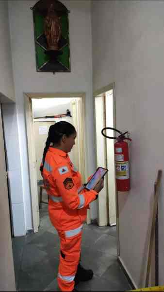 Bombeiro militar em traje de segurança laranja utilizando um tablet para inspeção em um corredor com um extintor de incêndio visível ao fundo.