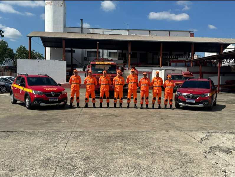 Equipe do 10º Batalhão de Bombeiros Militar em uniformes laranja, alinhada em frente a viaturas e instalações do quartel.