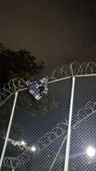 Parte de uma cerca de arame farpado com uma roupa pendurada no topo, sob um céu noturno.