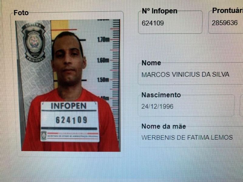 Foto de um detento com a ficha de identificação, usando uma camisa vermelha, posando em frente a uma parede com marcações de altura e um emblema da polícia.