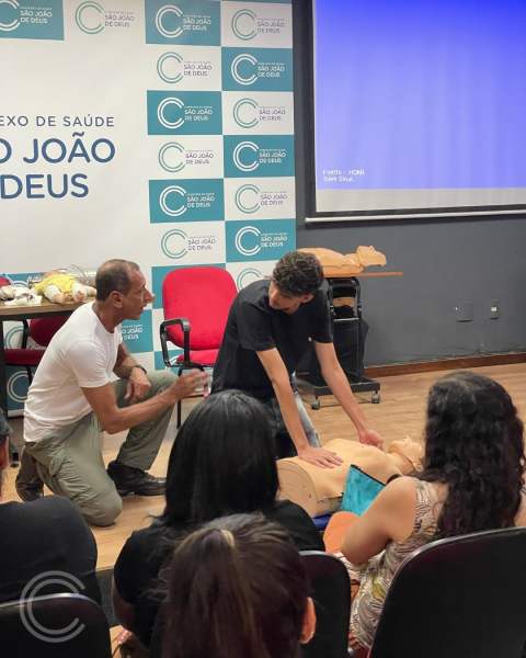 Um instrutor demonstra técnicas de primeiros socorros a um aluno utilizando um manequim em uma sala de aula da Escola Técnica São João de Deus, com outros participantes assistindo.