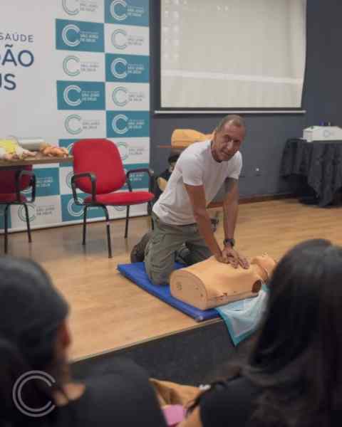 Instrutor demonstrando técnicas de reanimação em um boneco durante um aulão de primeiros socorros na Escola Técnica São João de Deus, com alunos observando ao fundo.