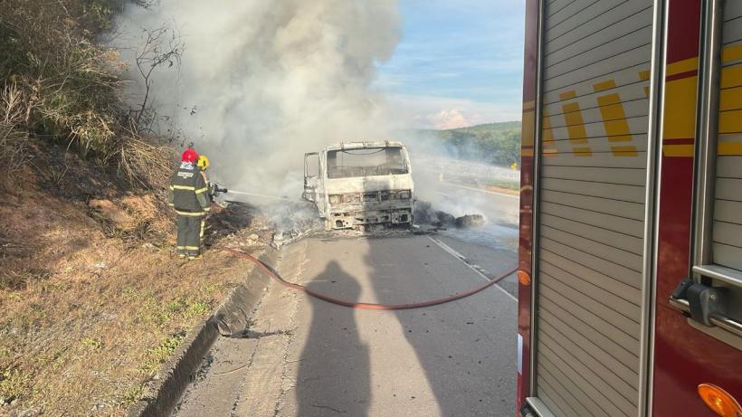 Bombeiros combatendo incêndio em caminhão na BR 262, com fumaça e chamas visíveis, ao lado de uma viatura do Corpo de Bombeiros.
