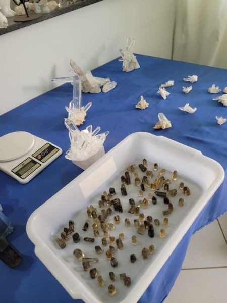 Um recipiente com cristais de quartzo em diferentes tamanhos e cores, colocado sobre uma mesa azul, ao lado de uma balança de precisão e vários cristais dispostos ao redor.