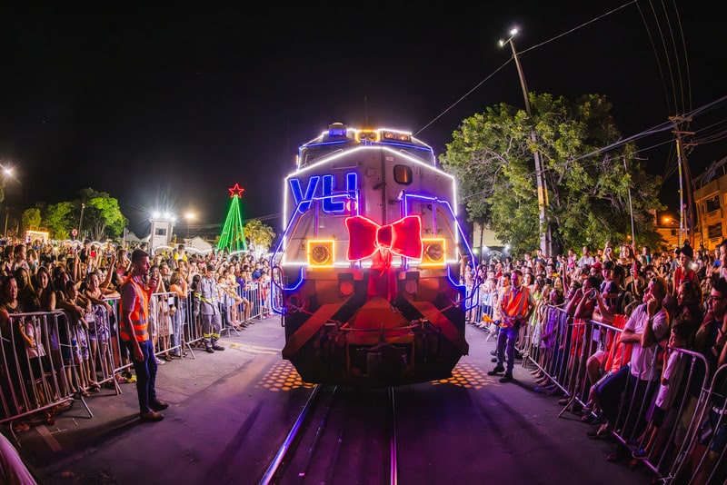 Locomotiva VLI decorada com luzes e laço vermelho, parada em frente a uma multidão em um evento natalino à noite.
