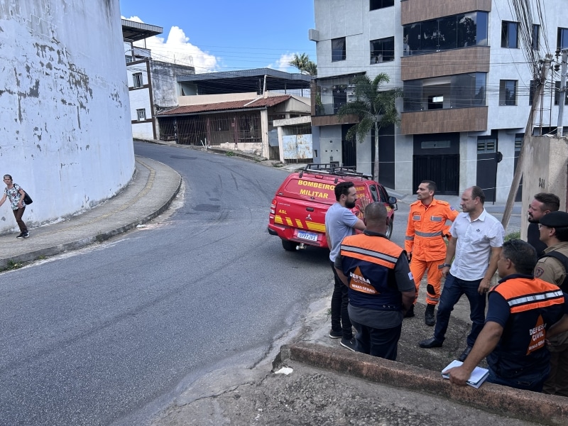 Grupo de profissionais, incluindo integrantes da Defesa Civil e Bombeiros, discutindo medidas de segurança em uma rua próxima a um muro em risco de desabamento.