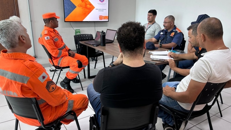 Reunião de profissionais em um ambiente interno, discutindo a simulação de emergência para o período chuvoso. No centro, um membro em uniforme laranja, cercado por outros participantes em cadeiras, com um projetor exibindo informações na parede.