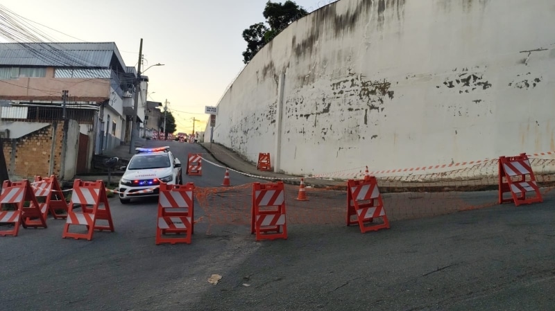 Barricadas de segurança fechando o acesso à Rua 10 de Julho, com um veículo da polícia estacionado próximo ao muro visivelmente danificado em Divinópolis.