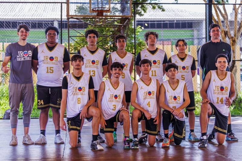 Jogadores da equipe JR09 Basketball posam juntos em quadra, exibindo camisetas brancas com detalhes em preto e dourado, após uma competição na Liga Metropolitana de Basquete de Minas Gerais.