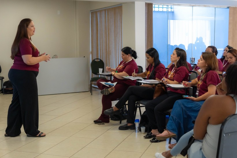 Uma instrutora apresenta um conteúdo a um grupo de mulheres participantes em um ambiente de capacitação, todos usando camisetas vermelhas e em atenção à palestra.