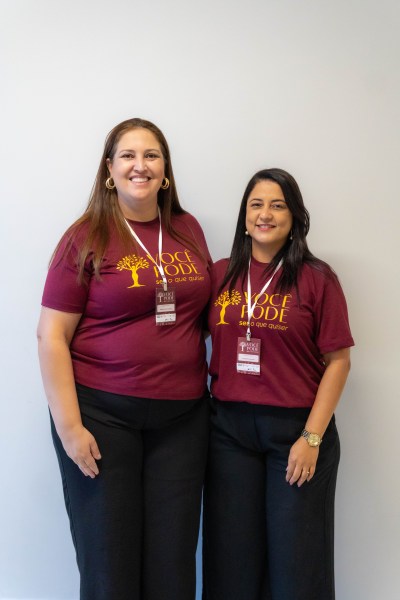 Duas mulheres sorridentes vestindo camisetas do projeto 'Você Pode', posando em um ambiente interno com parede clara.