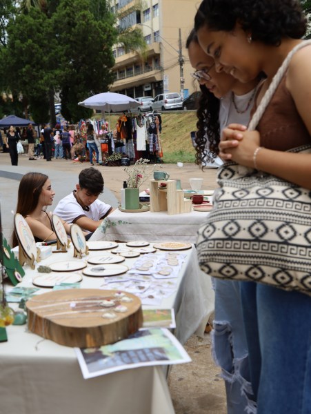 Três pessoas admirando produtos artesanais em uma banca durante a FEIRA ASTRAL, com expositores ao fundo e um ambiente vibrante.