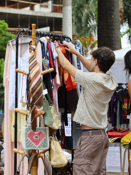 Pessoa examinando roupas penduradas em um cabideiro durante a Feira Astral, com um ambiente de feira ao fundo.