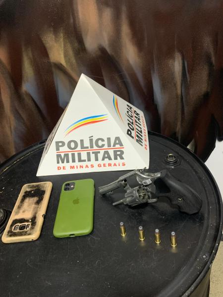 Mesa com uma arma de fogo, um celular, um case de celular e cápsulas de munição, com um cone de sinalização da Polícia Militar de Minas Gerais ao fundo.
