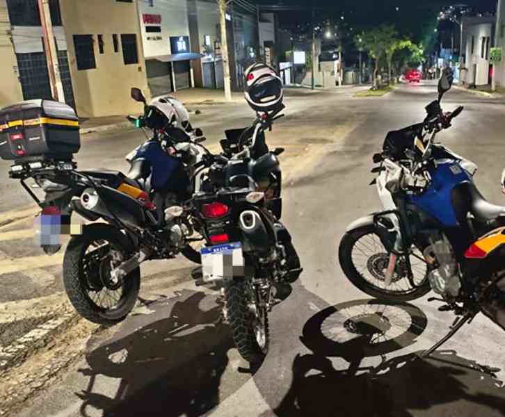 Imagem noturna de motocicletas da polícia estacionadas em uma rua, com sinais de patrulhamento e ação de segurança.