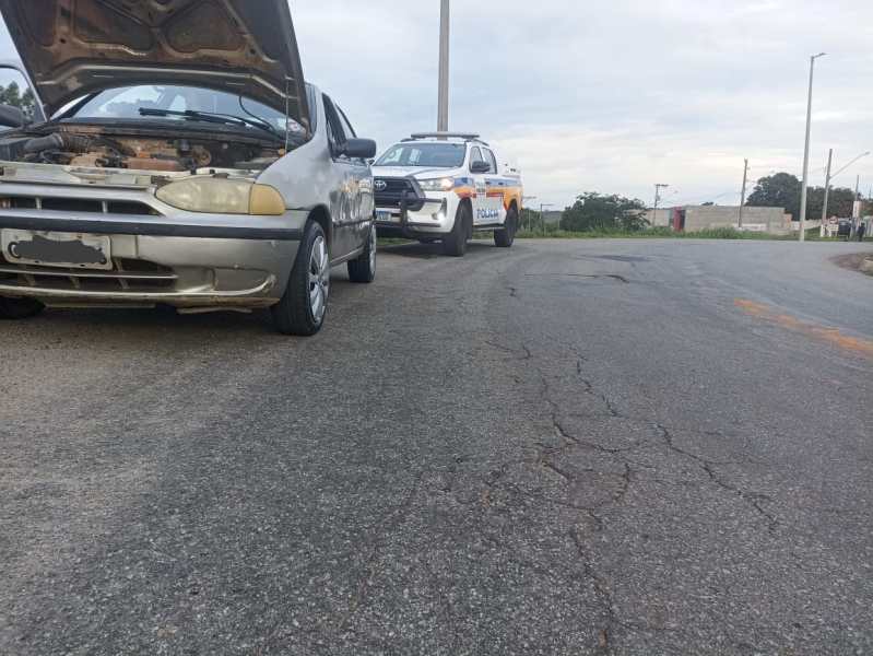 Um Fiat Palio com o capô aberto estacionado à beira da estrada, ao lado de uma viatura policial.