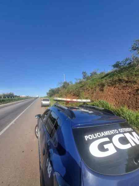Imagem de um carro da Guarda Civil Municipal estacionado à beira da rodovia, com uma estrada e vegetação ao fundo.