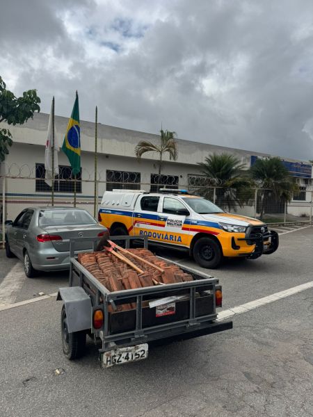 Um veículo da Polícia Militar Rodoviária estacionado em frente a um edifício, com um carro cinza e um semi-reboque carregado de materiais ao lado.