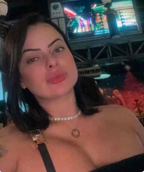 Close-up of a woman with long dark hair, wearing a black off-shoulder top and a necklace, posing in an indoor setting with colorful lights in the background.