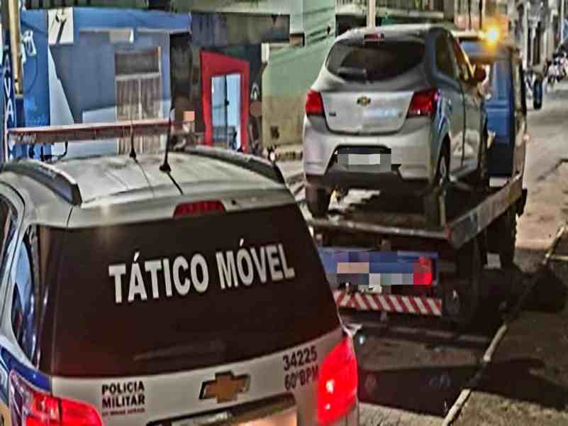 Veículo sendo rebocado por uma caminhonete da Polícia Militar em uma rua à noite, com outros veículos e construções ao fundo.