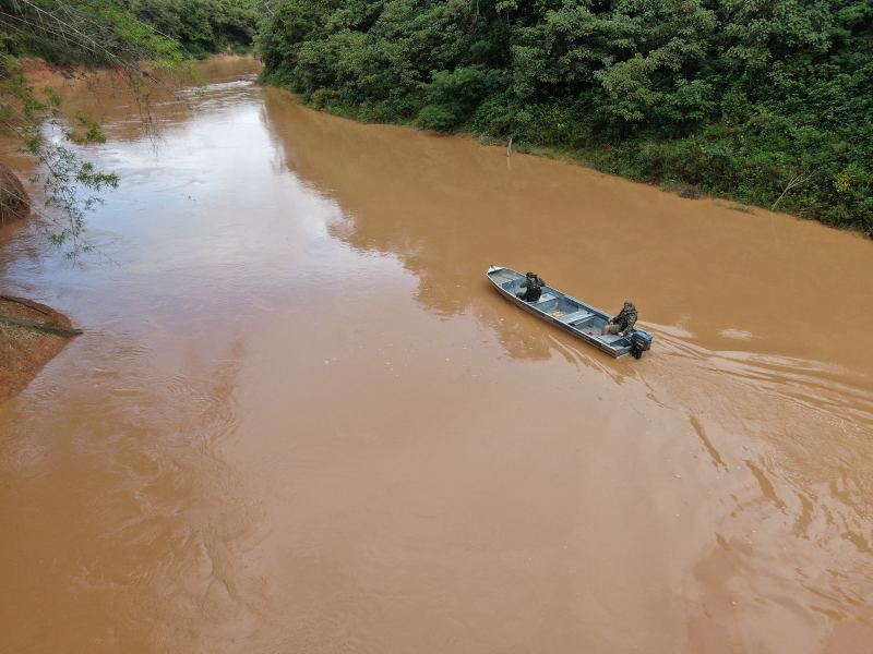 Um barco com dois homens da Polícia Militar de Meio Ambiente navegando em um rio de água barrenta, cercado por vegetação densa.