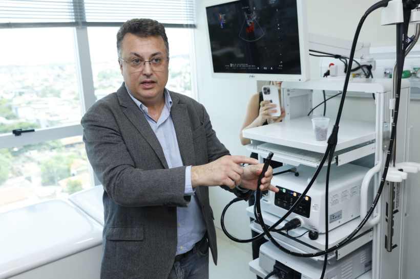 Um médico explicando o funcionamento de um aparelho de endoscopia em um ambiente clínico moderno, com um monitor ao fundo exibindo imagens.