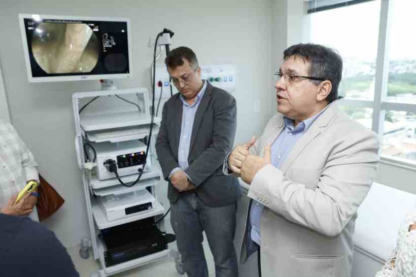 Dois homens em um ambiente médico, um falando enquanto o outro escuta, com um equipamento de endoscopia visível ao fundo e uma tela mostrando uma imagem endoscópica.