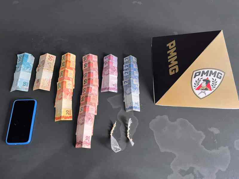 Um celular, uma variedade de notas em dinheiro e duas pequenas porções de droga sobre uma superfície escura, ao lado de uma caixa com logo da Polícia Militar de Minas Gerais.