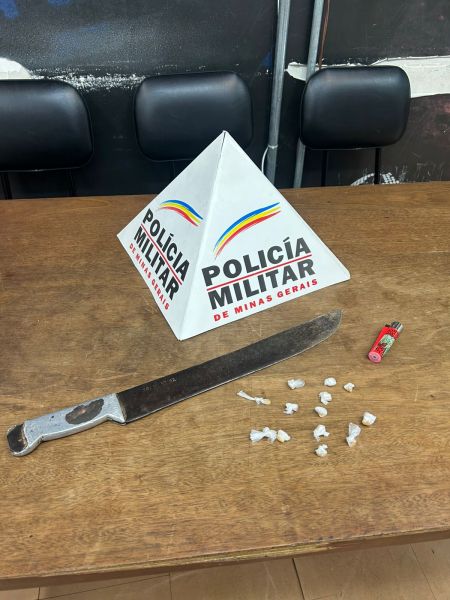 Uma mesa com uma placa da Polícia Militar de Minas Gerais, um facão, um isqueiro e pedras de substância semelhante ao crack dispostas ao lado.